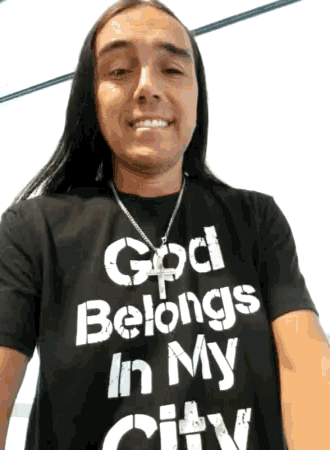 a man with long hair wears a black shirt that says god belongs in my city