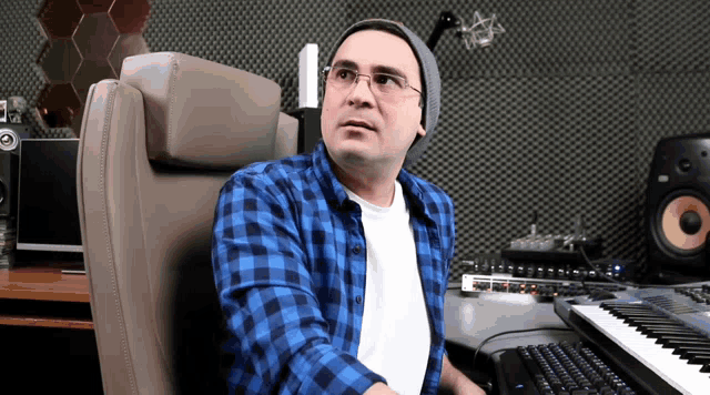 a man wearing glasses and a beanie sits in front of a keyboard and speakers