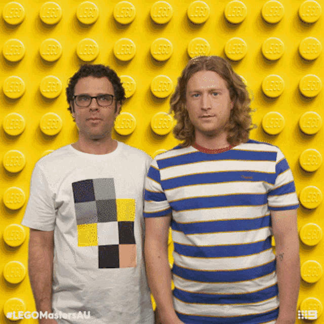 two men standing in front of a yellow wall with lego bricks on it