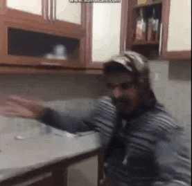 a man is standing in a kitchen with his arms outstretched and wearing a hat .