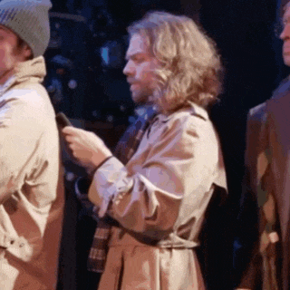 a man in a trench coat is holding a cell phone while standing next to another man .