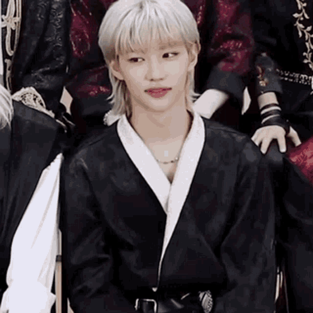 a young man with blonde hair is wearing a black jacket and a necklace .