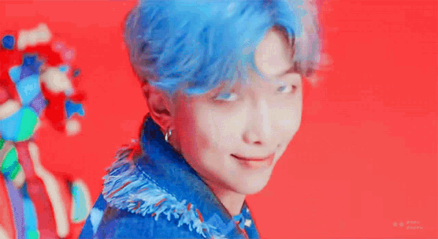 a young man with blue hair is standing in front of a red background and making a funny face .