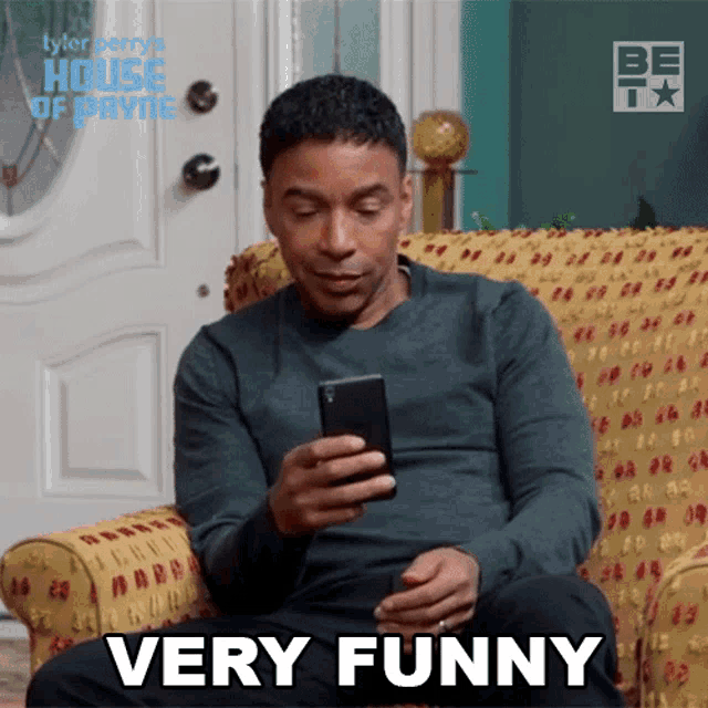a man is sitting on a couch looking at his cell phone and says very funny