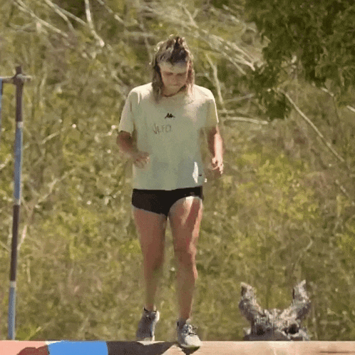 a woman in a white shirt and black shorts is jumping over a wooden beam .