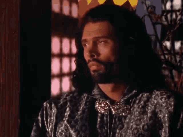 a man with long hair and a beard is standing in front of a stained glass window in a room .