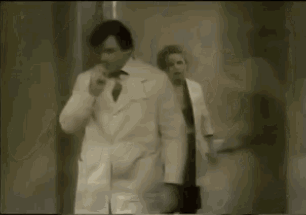 a man in a white coat and tie is walking down a hallway next to a woman in a white coat .