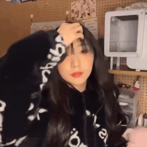 a girl in a black hoodie is standing in a room holding her head .