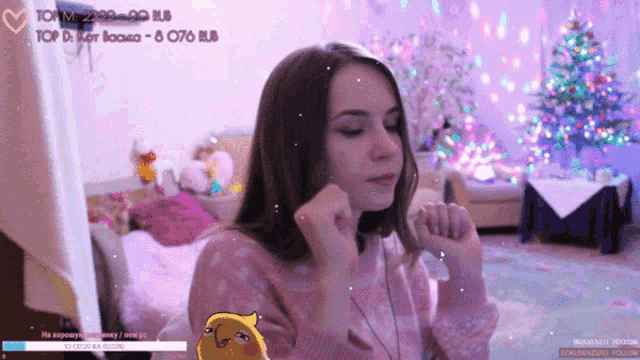 a girl in a pink sweater is standing in front of a christmas tree with a yellow bird on her shoulder