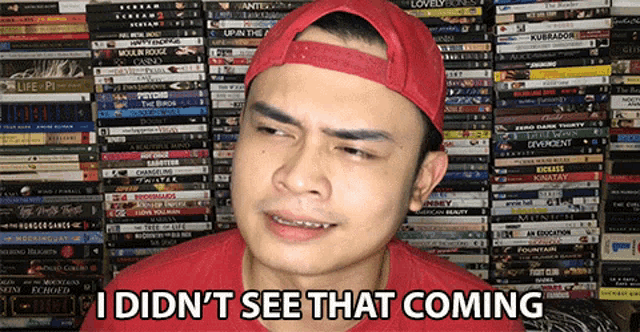 a man wearing a red hat says " i did n't see that coming " in front of a wall of dvds