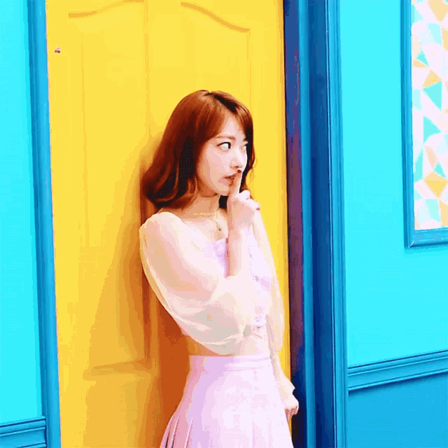 a woman in a pink dress is standing in front of a yellow door holding her finger to her mouth .