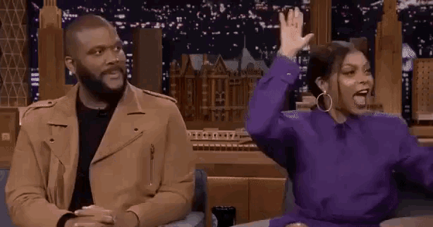 a man and a woman are sitting next to each other on a couch and the woman is raising her hand .