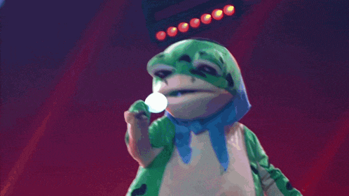 a green and white frog with a blue scarf around its neck is on a stage