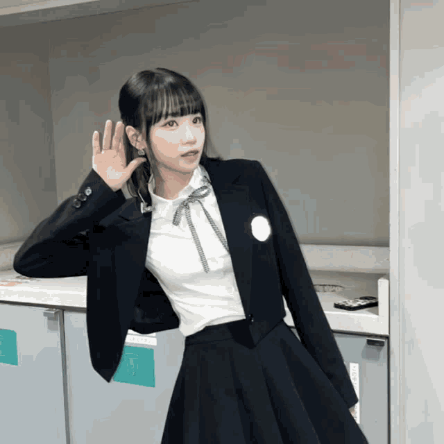 a girl in a school uniform holds her hand to her ear while wearing a jacket