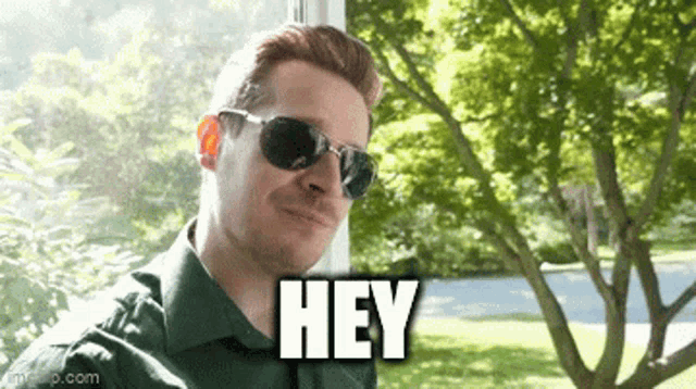 a man wearing sunglasses and a green shirt is standing in front of a window and says hey .