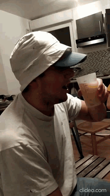 a man wearing a hat and a white shirt is drinking from a measuring cup