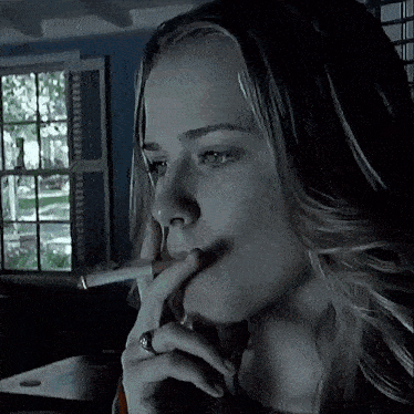 a woman is smoking a cigarette in a dark room with a window in the background .