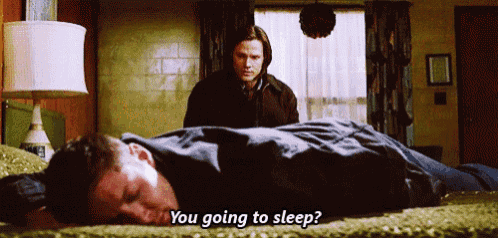 a man is laying on a bed while another man stands behind him and says " you going to sleep ? "