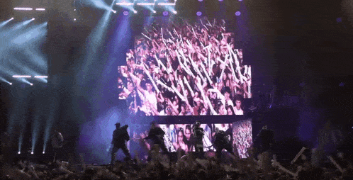 a crowd of people are dancing on a stage in front of a large screen