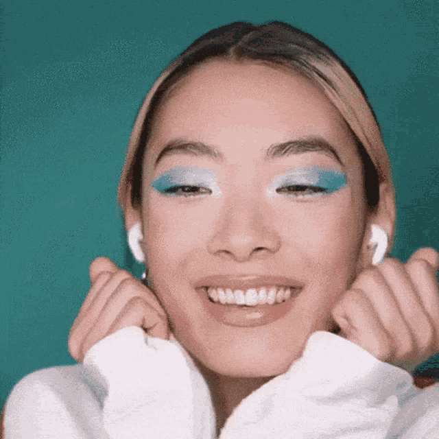 a woman wearing blue eyeshadow and white ear buds is smiling and making a funny face .