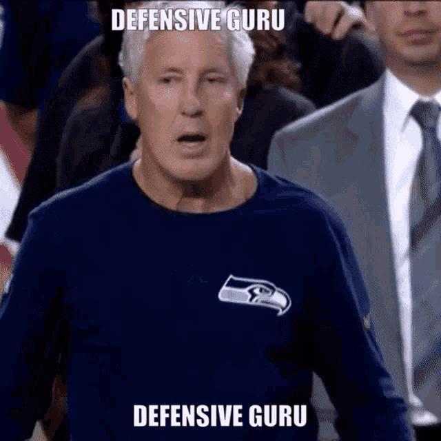 a man wearing a seahawks shirt stands in front of a crowd and says " defensive guru "
