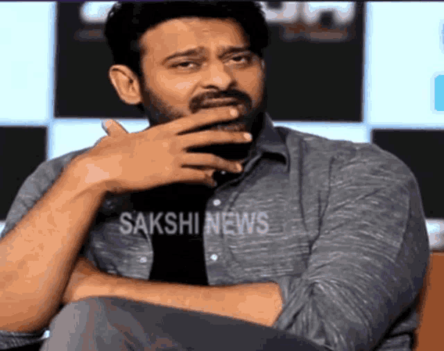 a man with a beard is covering his mouth with his hand while wearing a sakshi news shirt .