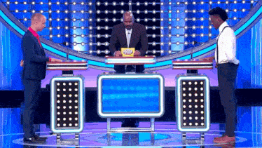 a man in a suit is sitting at a table on a game show with two other people .