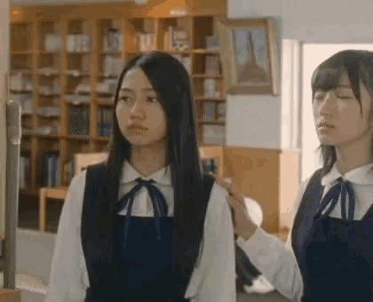 two girls are standing next to each other in a library and one of them is putting her hand on the other 's shoulder .