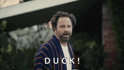 a man in a robe is standing in front of a house and says duck !