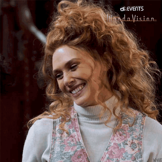 a woman with curly red hair is smiling and wearing a floral vest .