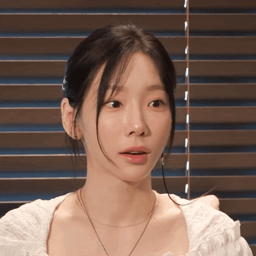 a woman wearing a white top and a necklace looks at the camera with a surprised look on her face