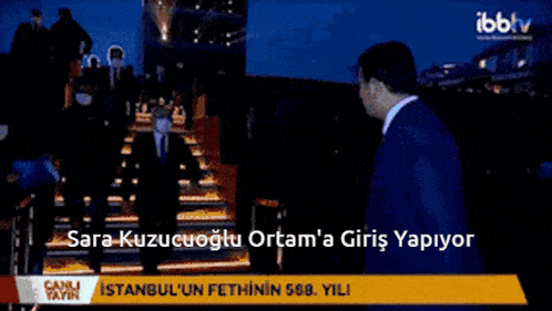 two men in suits and ties are walking in front of a sign that says ' sara kuzucuoglu ortam ' on it