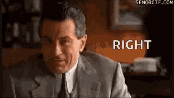 a man in a suit and tie is sitting at a desk and looking at the camera with the word right above him .
