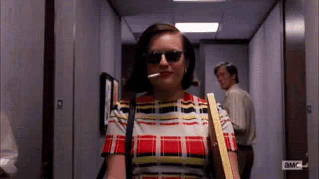 a woman wearing sunglasses and a plaid shirt is walking down a hallway with a man behind her ..