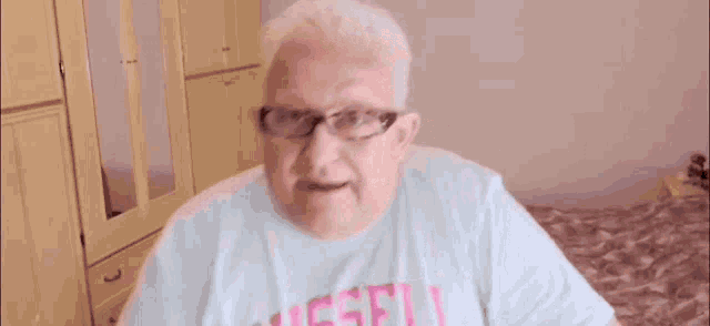 an elderly man wearing glasses and a blue shirt is standing in a bedroom .