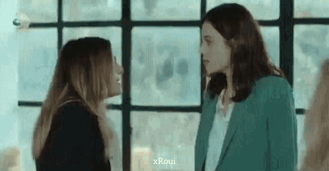two women are standing next to each other and looking at each other in front of a window .