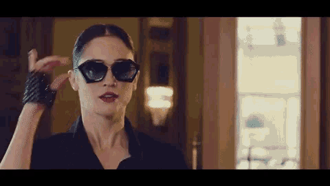 a woman wearing sunglasses and a black jacket stands in front of a window .