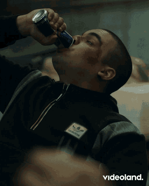 a man wearing a black adidas jacket drinks a can of soda