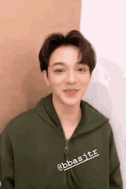 a young man wearing a green hoodie is standing in front of a wall and smiling .