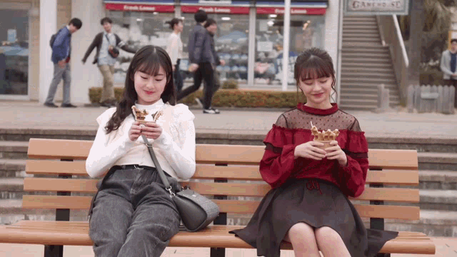 two girls are sitting on a bench eating sandwiches in front of a store that says bakery st.
