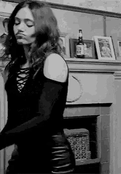 a woman in a black dress is standing in front of a fireplace in a black and white photo .