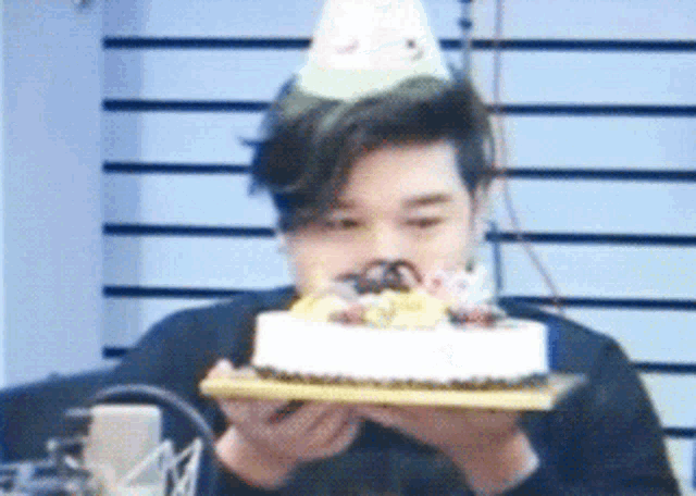 a man wearing a party hat holds a cake in front of his face