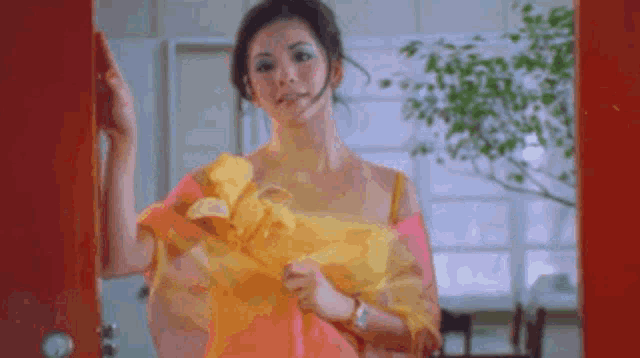 a woman in a pink and yellow dress is standing in front of a red door holding a yellow flower .