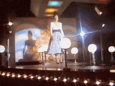 a woman in a dress is walking on a stage with lights behind her