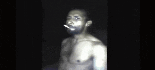 a man without a shirt is standing in the dark with his hands behind his head .