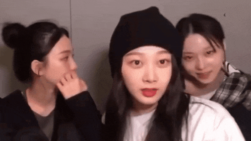 a group of three girls are standing next to each other . one of the girls is wearing a black hat .