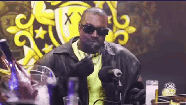 a man wearing sunglasses is talking into a microphone while sitting at a table .