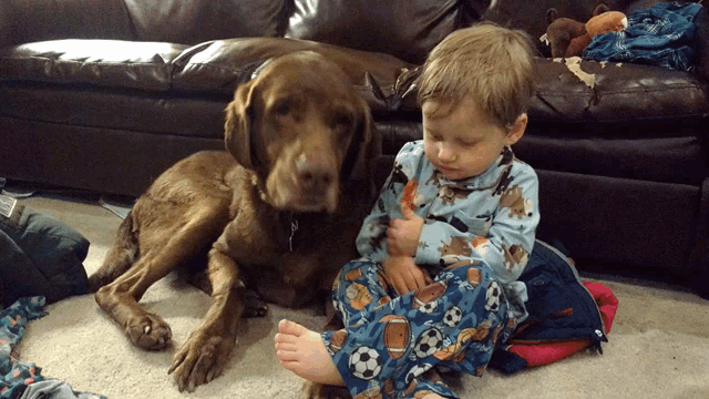 a little boy sitting on the floor next to a brown dog wearing football pajamas