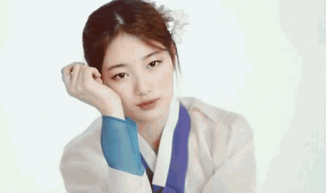 a woman in a traditional korean dress with a flower in her hair is looking at the camera with her hand on her face .