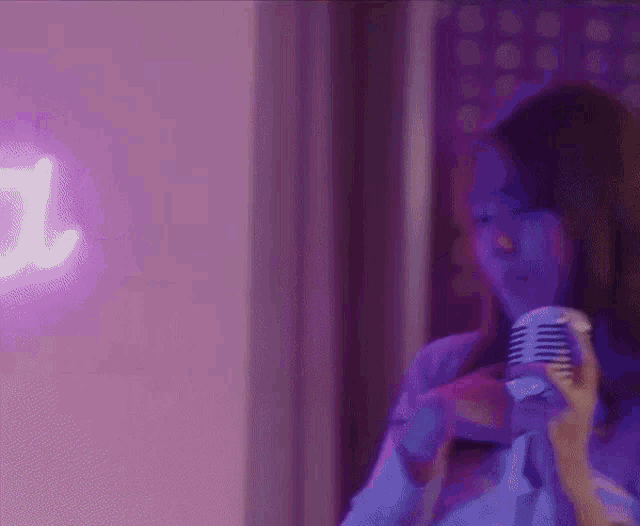 a woman is singing into a microphone in front of a purple neon sign .